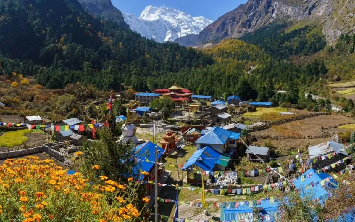 Colorful Himalayan village with blue-roofed houses, prayer flags, and Mount Manaslu backdrop