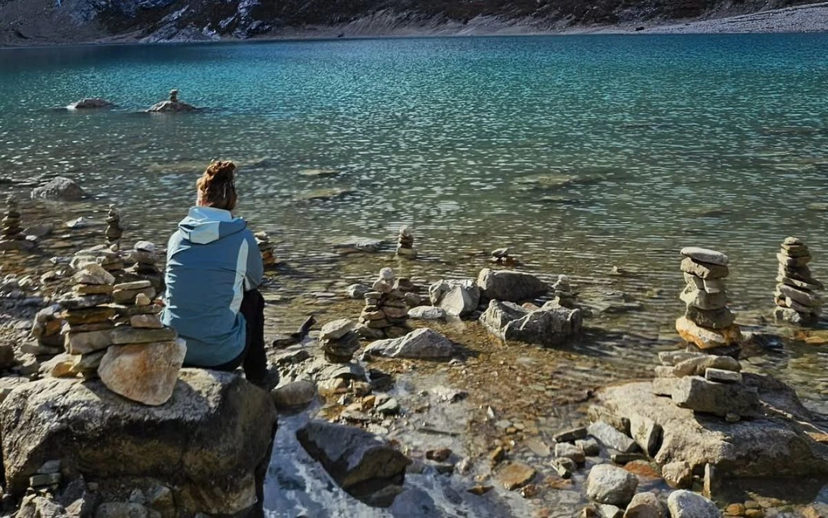 Person sitting by turquoise mountain lake surrounded by stacked stone cairns