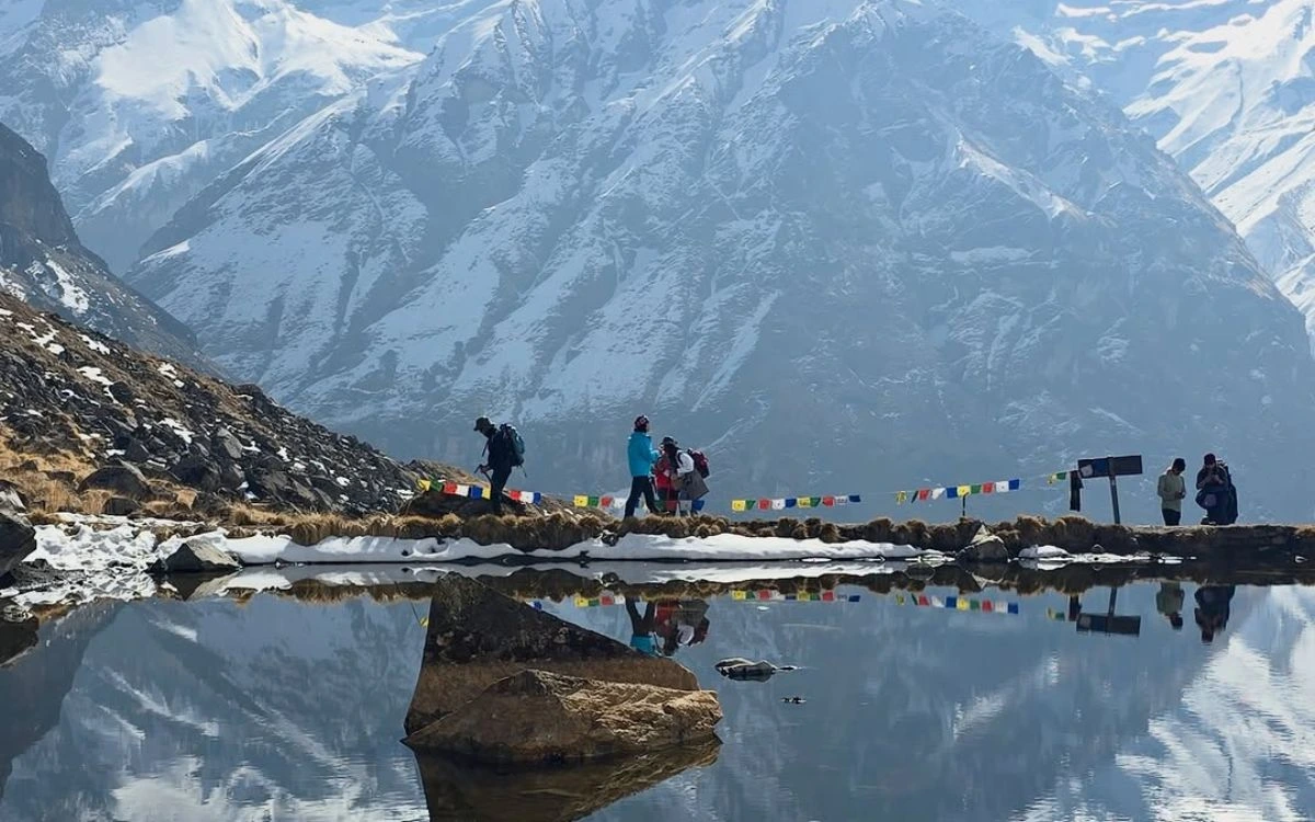 Trekkers walking beside a still mountain lake reflecting snow peaks and prayer flags at ABC
