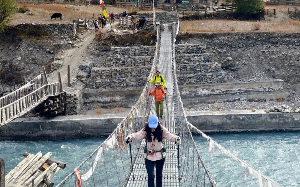 Trekkers crossing suspension bridge over turquoise glacial river in Himalayan village along Annapurna Circuit