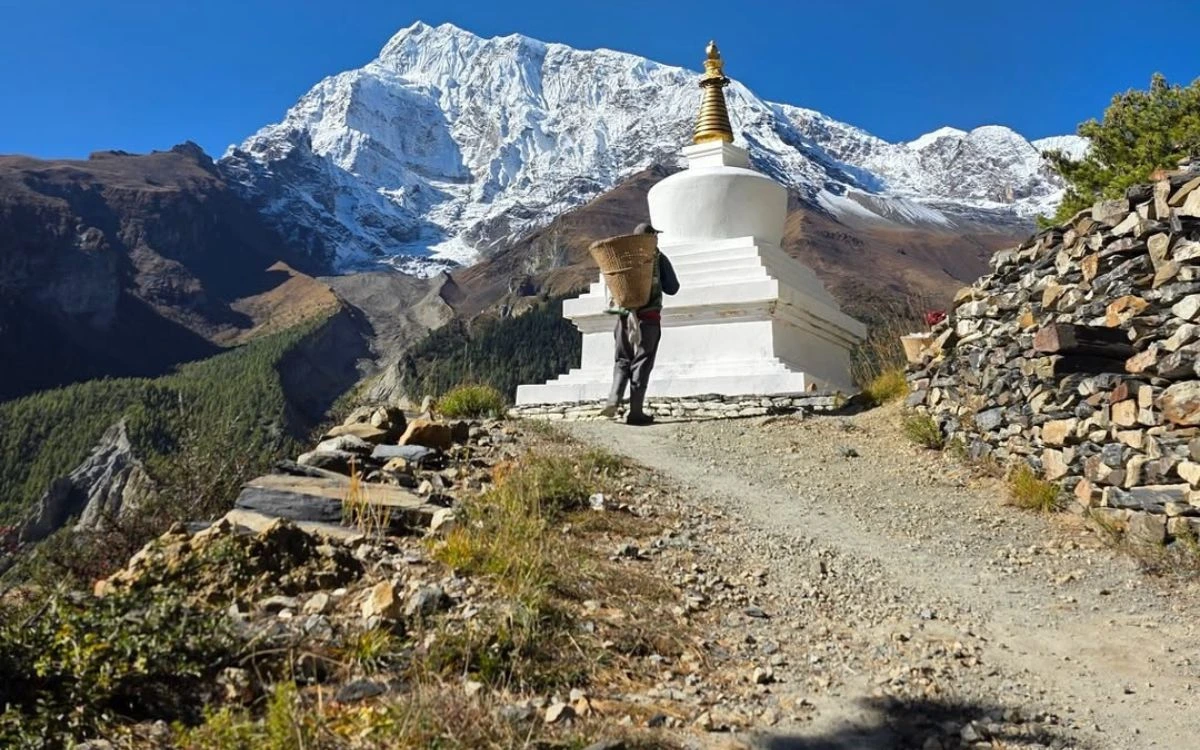 A porter carrying a doko basket past a white Buddhist stupa on the Annapurna Circuit trail