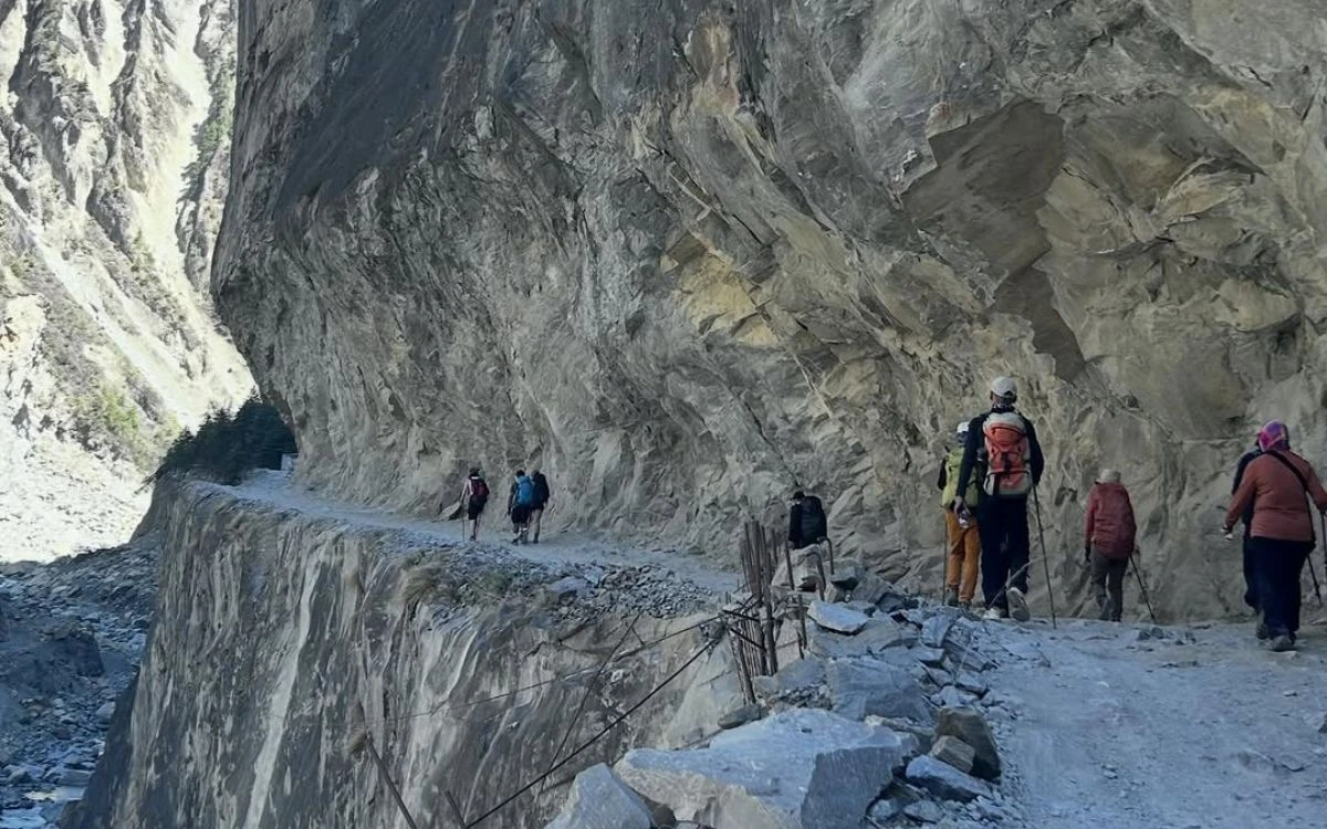 Trekkers walking single file along a cliff-edge road beneath overhanging rock walls on the Annapurna Circuit