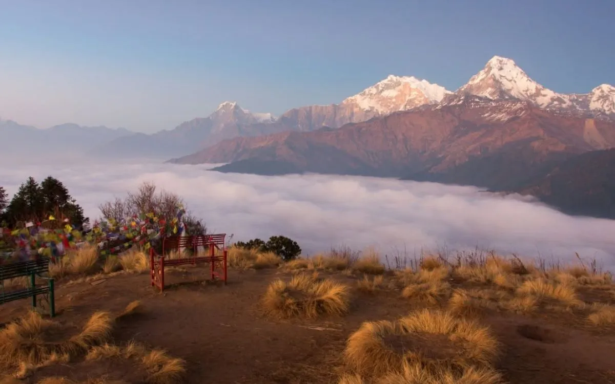 Mountain viewpoint with prayer flags and bench overlooking snow-peaked Annapurna range above sea of clouds