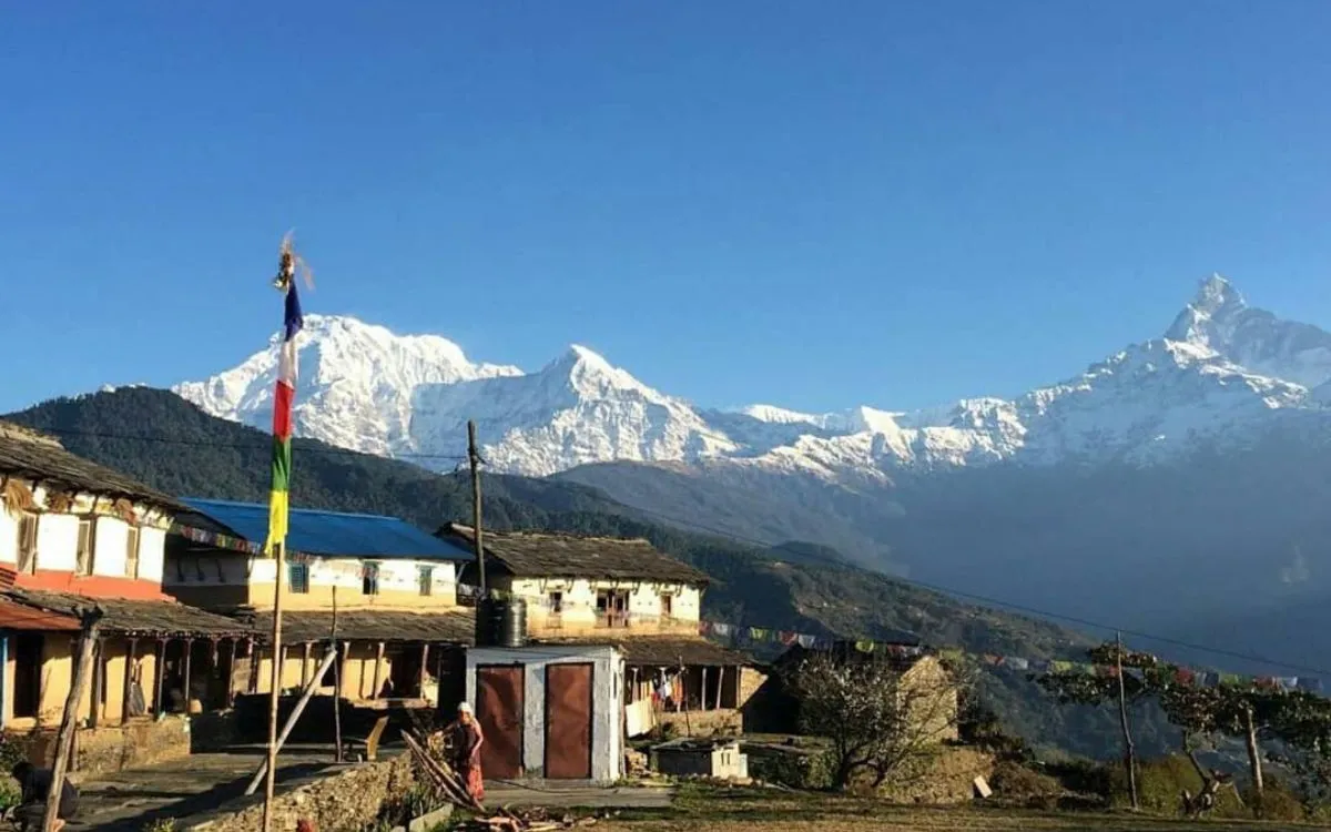 Traditional mountain village with stone houses and prayer flag pole, snow-capped Annapurna range visible in distance