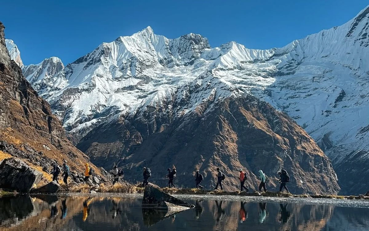 A group of trekkers walking in a line beside a still reflective lake with massive snow-capped peaks behind