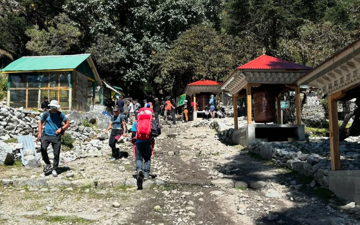 Trekkers passing through a mountain village checkpoint with a traditional prayer wheel and teahouse along a Nepal trail