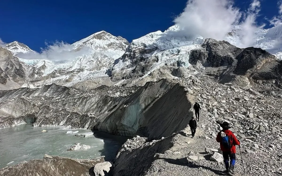  Trekkers walking along rocky terrain near a glacial lake with snow-capped Himalayan peaks in the background.