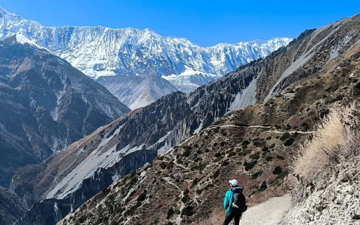 A solo trekker on a narrow high-altitude trail with a dramatic Annapurna valley and snow-covered peaks
