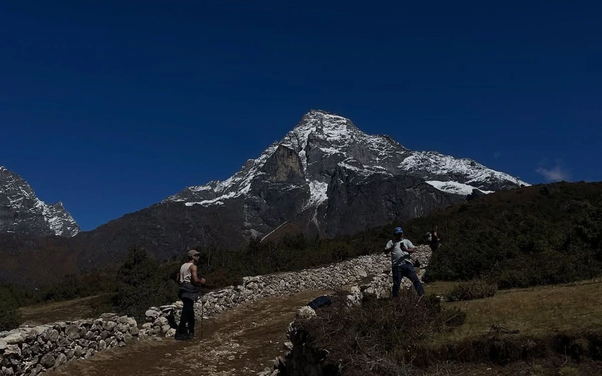 Trekkers hiking a stone-walled trail near Khumjung with a dramatic snow-dusted peak dominating the skyline