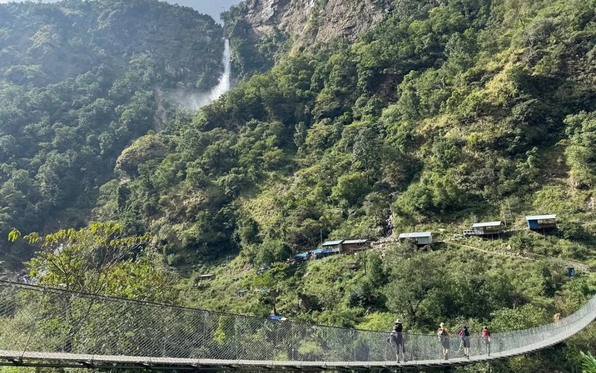 Trekkers crossing suspension bridge over Budhi Gandaki River with waterfall and lush green hillsides