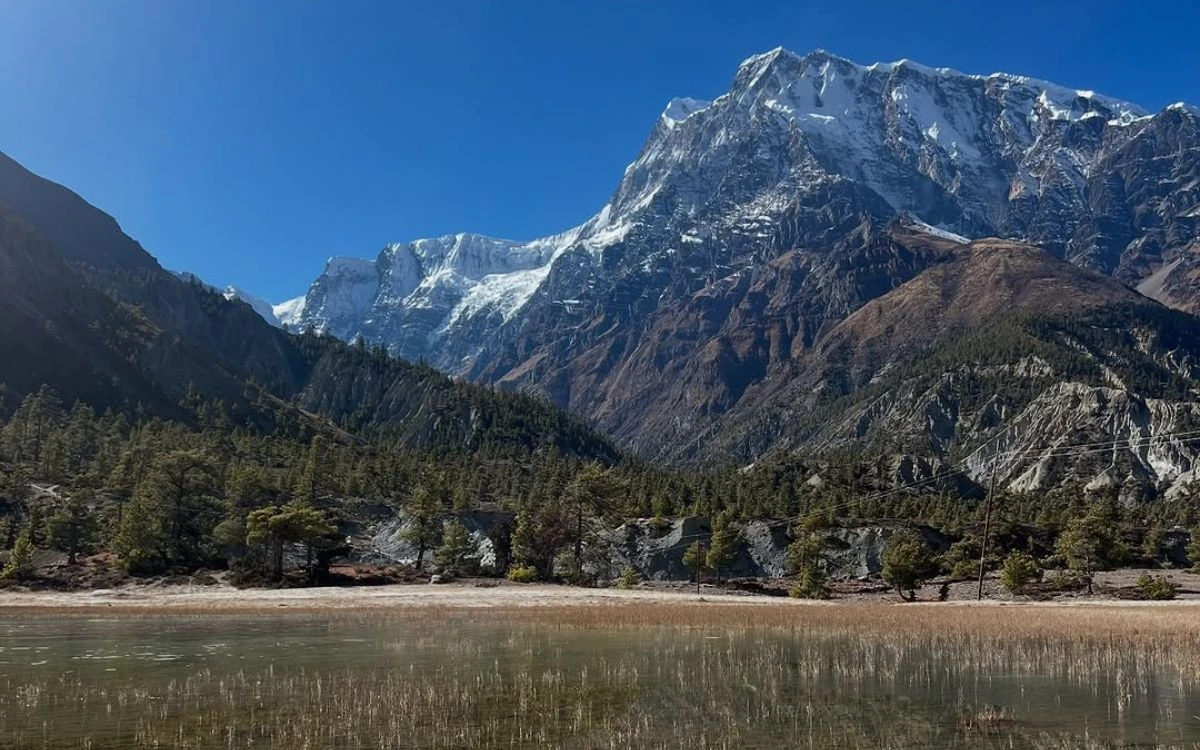 Calm lake reflecting pine trees and a sheer snow-capped peak at the entrance to Manang Valley