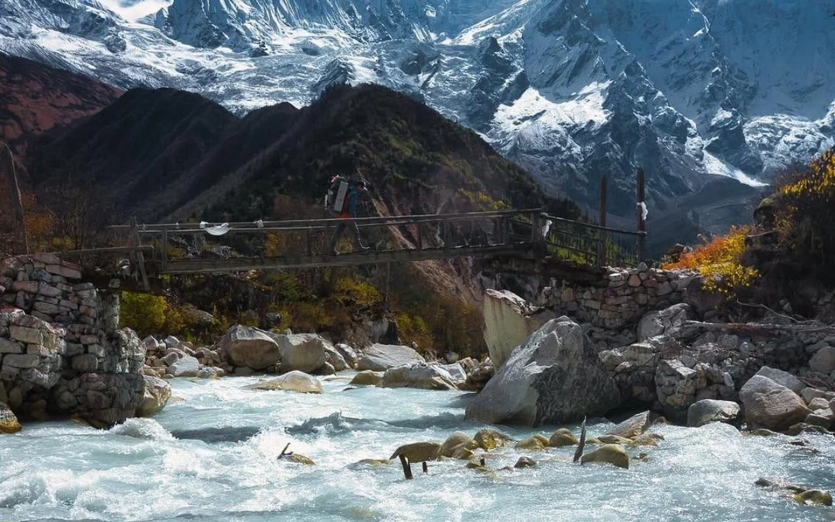Suspension bridge over rushing river with snow-capped Manaslu peaks behind