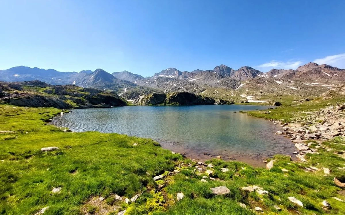 Calm alpine lake surrounded by green grass and rocky mountain ridges on a clear July day