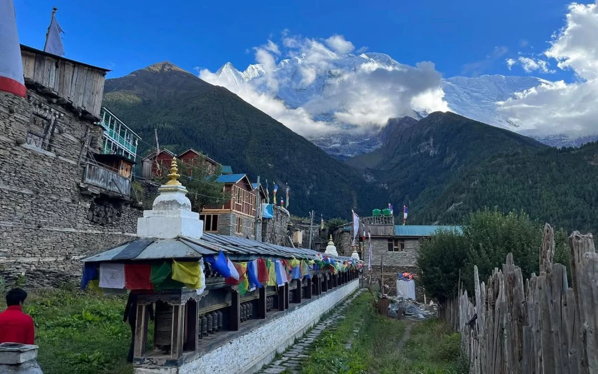 Colorful prayer flags and prayer wheels line a path through a Himalayan village with snow-capped peaks behind