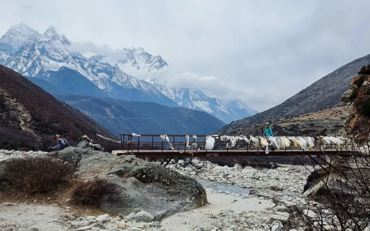Trekkers crossing a prayer-flag-draped bridge over a glacial stream with snow-capped Himalayan peaks behind