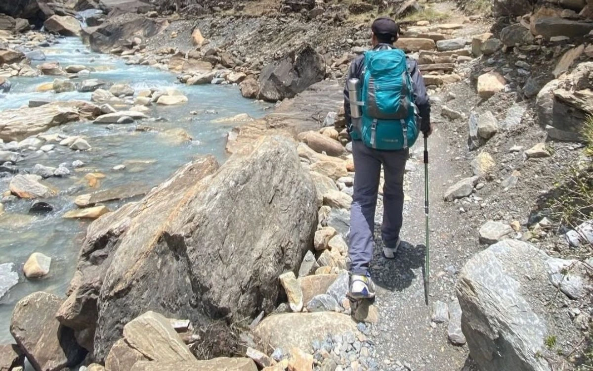A solo trekker with a blue backpack walking a rocky trail alongside a turquoise glacial river in Nepal