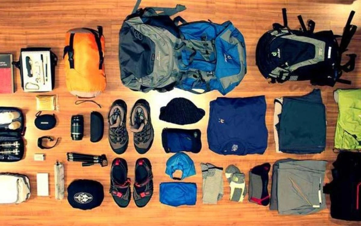 Flat lay photography of trekking equipment on wood including backpacks, hiking boots, sleeping bag, jackets, and accessories.
