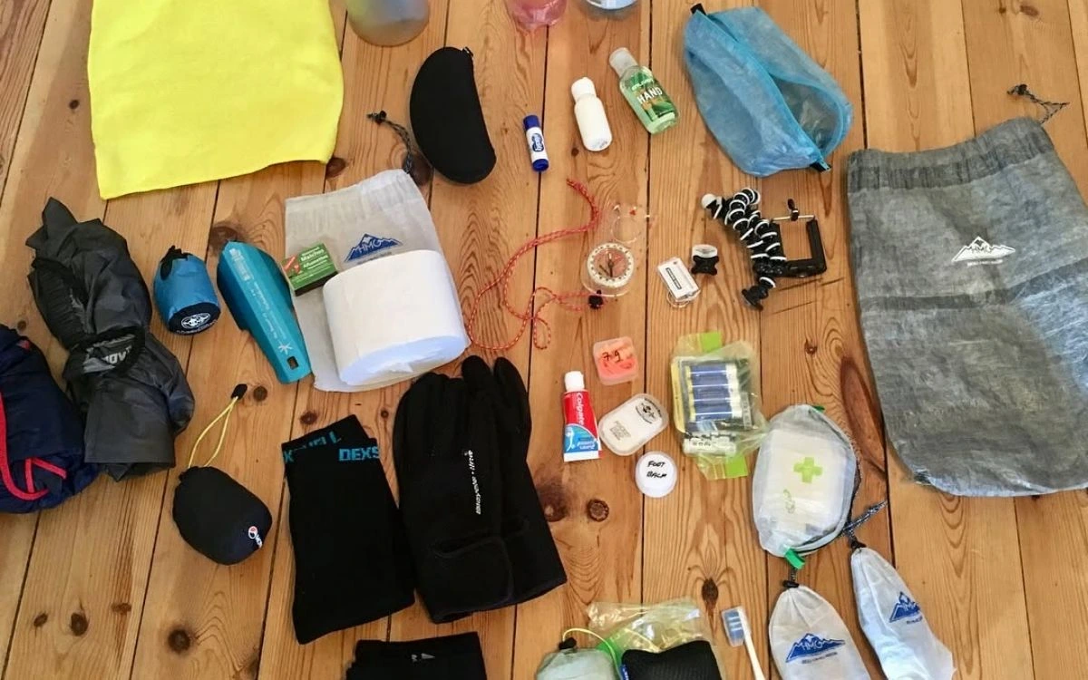 Trekking gear laid out on a wooden floor including backpack, sleeping bag, jackets, socks, water bottles, and accessories.