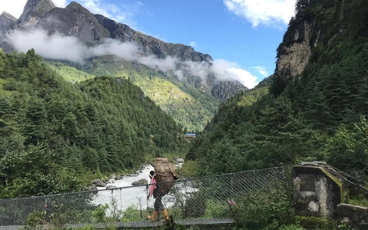 Porter with traditional basket crossing suspension bridge over glacial river in green valley