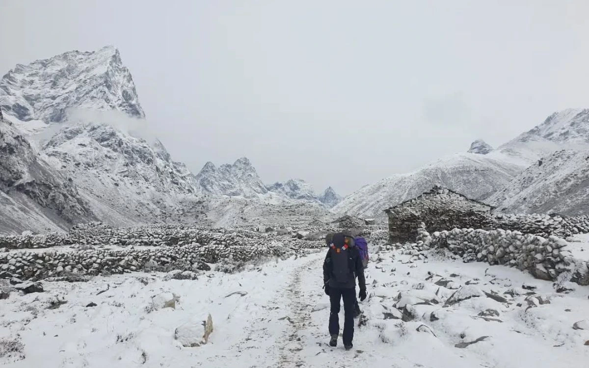 A trekker with a large backpack walking through a snowstorm past stone huts near Pheriche 