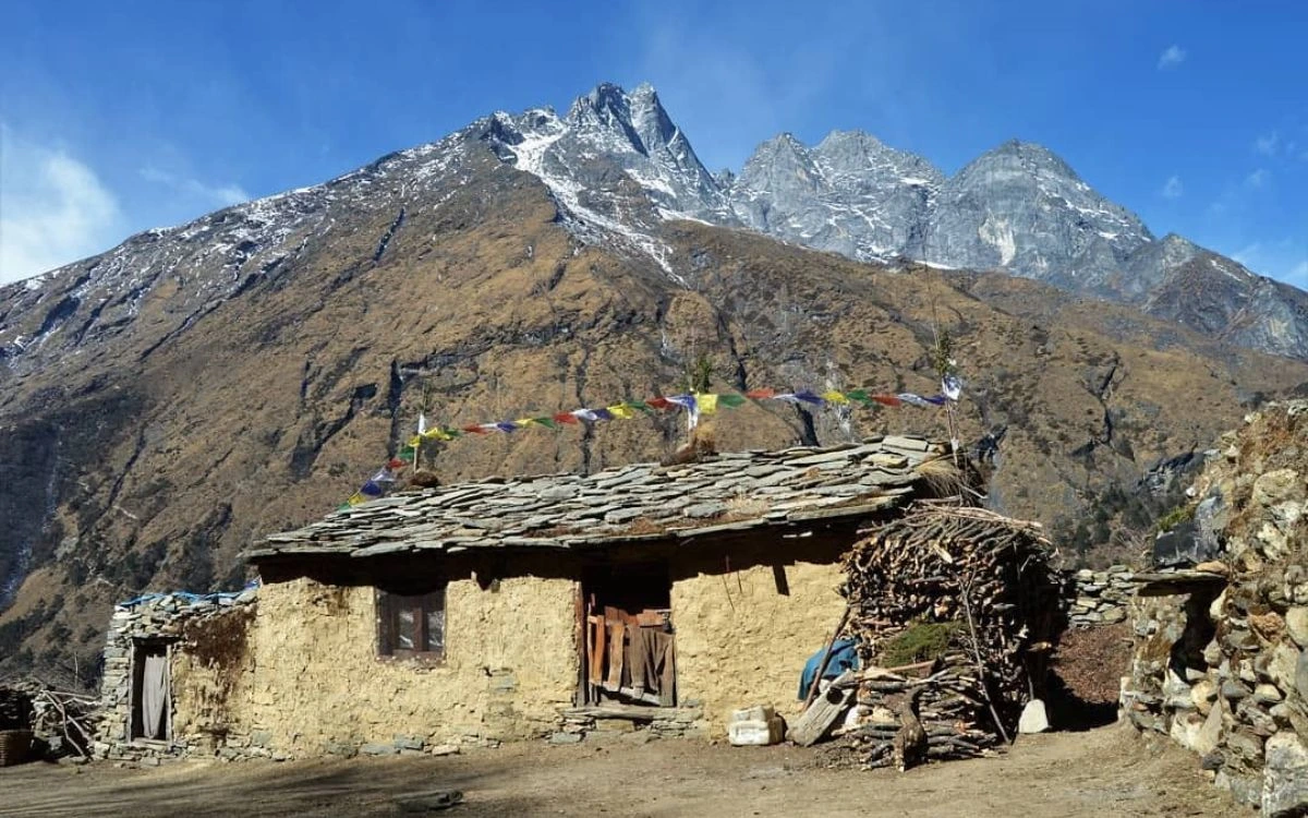 Traditional mud-and-stone Sherpa house with colorful prayer flags and a firewood stack against a rocky Himalayan ridge