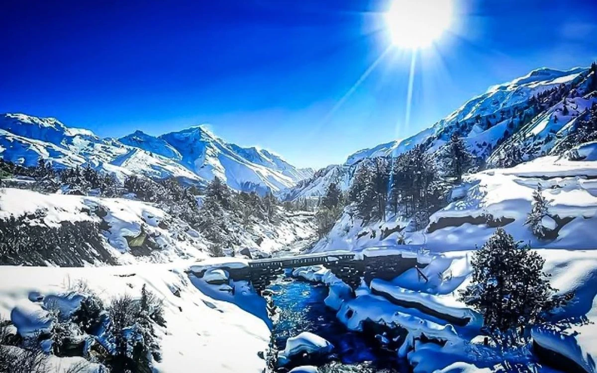 Snow-covered Himalayan valley near Pisang with a river, stone bridge, and pine trees under a bright sun