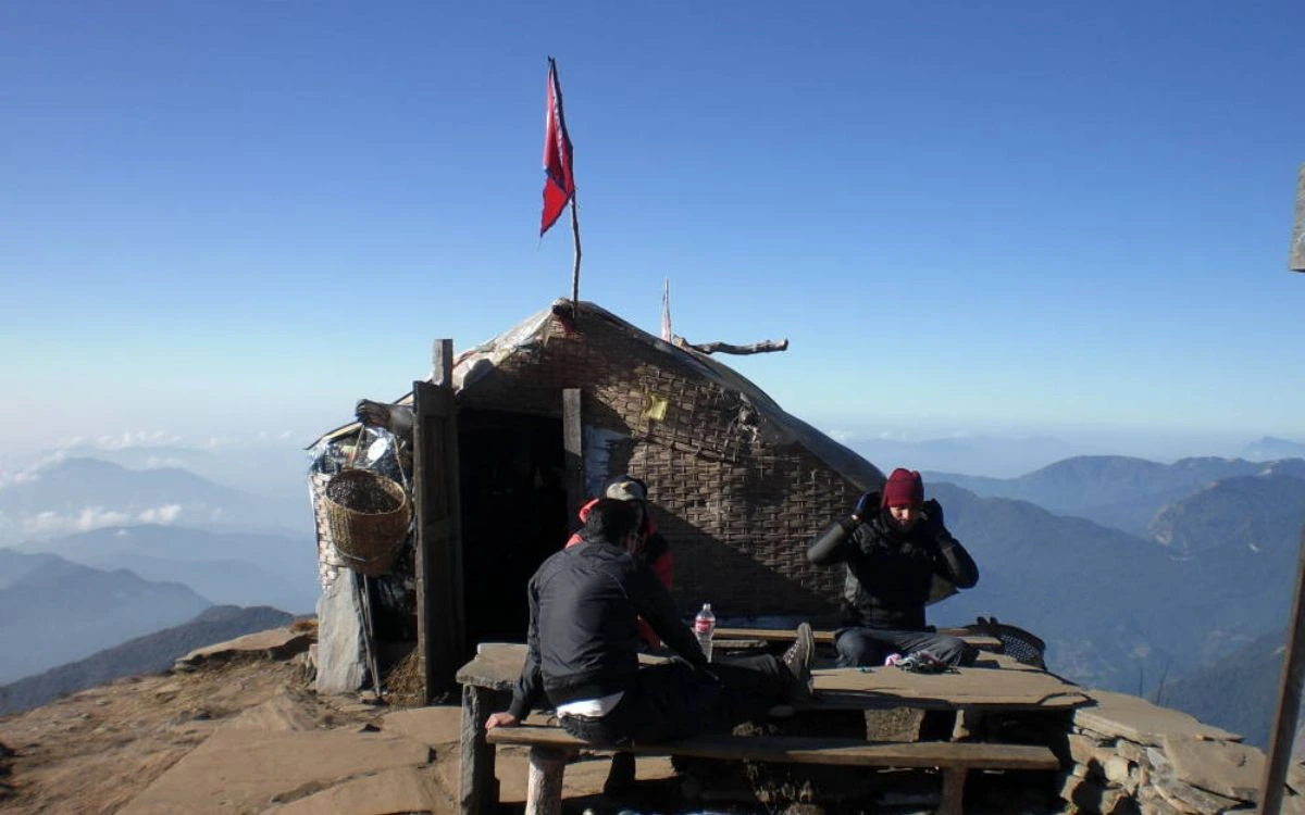 Trekkers resting at a rustic hilltop shelter with Nepali flag and panoramic mountain views