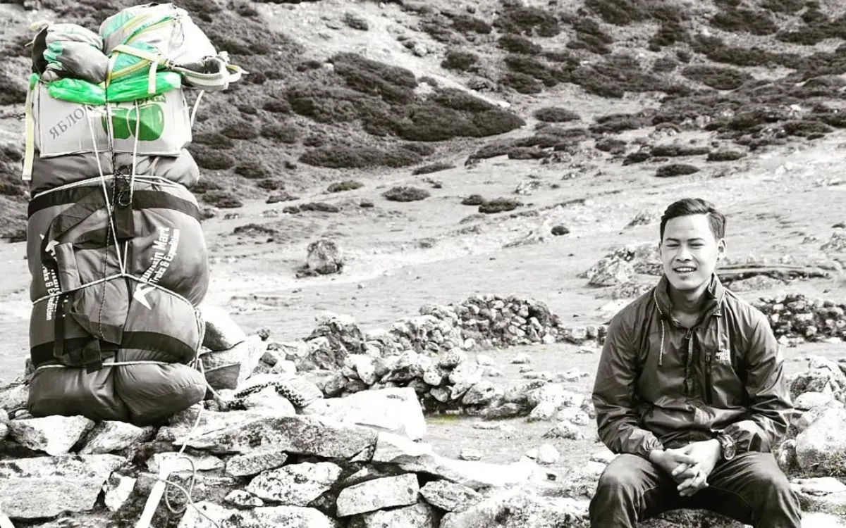 Sherpa guide resting beside heavy expedition pack near stone cairn in black and white