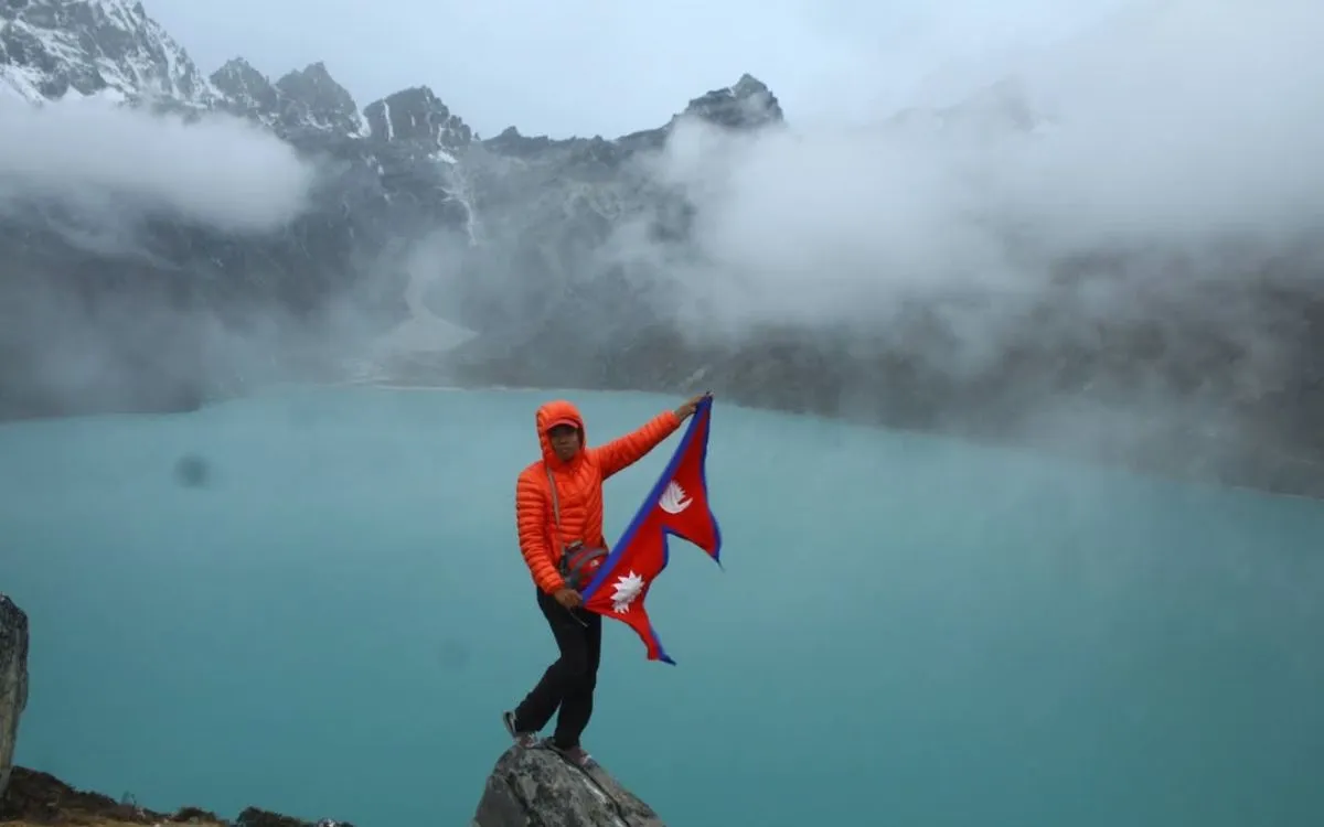 Hiker in orange jacket holding Nepal flag beside turquoise mountain lake