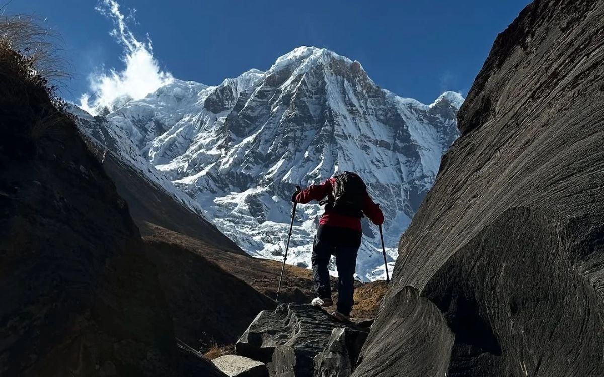 Solo trekker with poles climbing rocky steps toward a massive snow-capped Himalayan peak