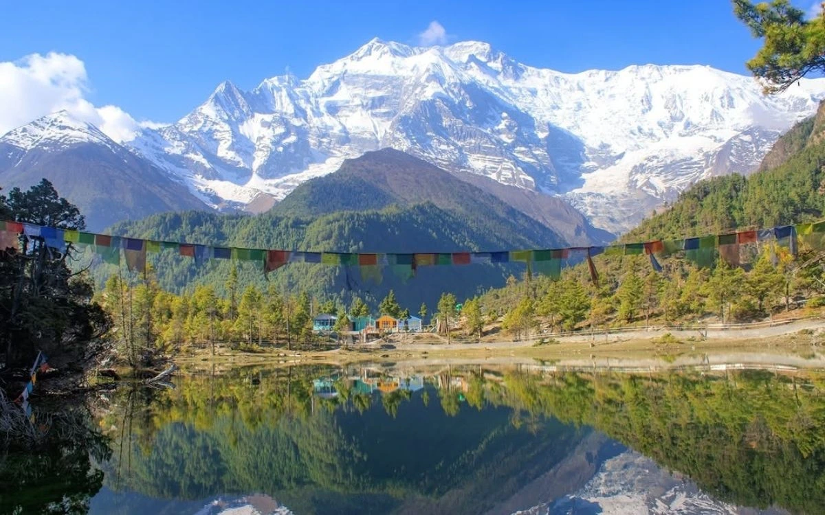 Prayer flags strung over a still lake reflecting Annapurna II and pine forest in the Annapurna Circuit