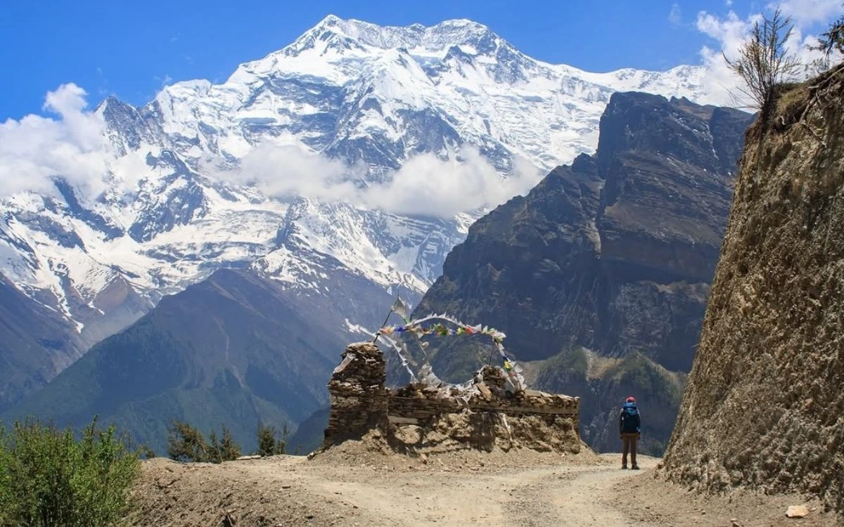 Trekker passing a stone mani wall with prayer flags and Annapurna massif towering above