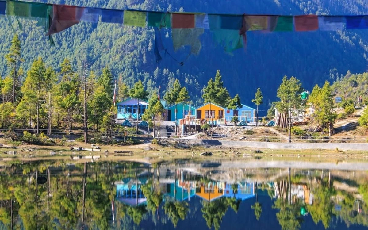 Colorful teahouse lodges reflected in a calm lake with pine forest and prayer flags above