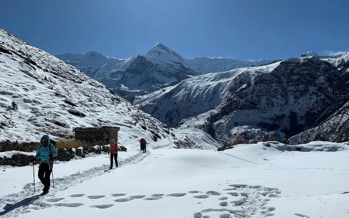 Trekkers hiking a snow-covered trail toward Thorong Phedi with a sharp mountain peak in the background