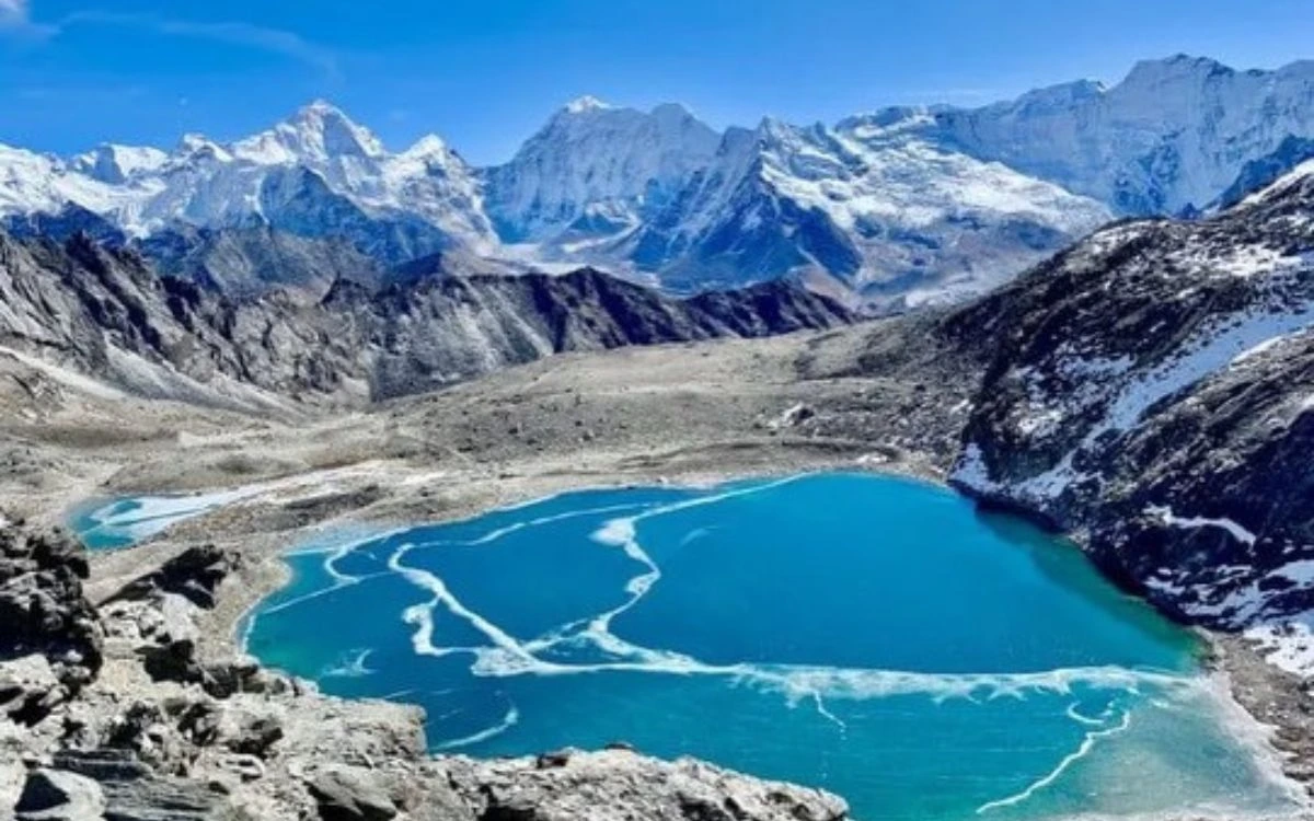 A bright blue glacial lake partially covered with patterns of white ice, surrounded by high snow peaks.