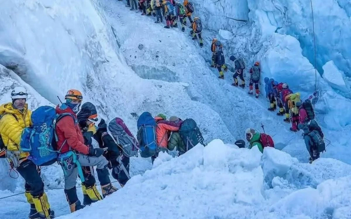 A long line of climbers wearing high-altitude gear waiting in a queue on a steep, snowy Everest slope. 