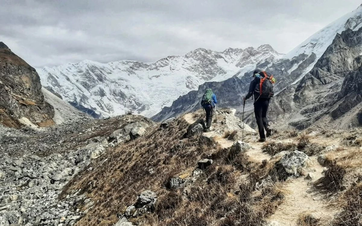 Two trekkers with backpacks and poles on a rocky high-altitude ridge approaching Kanchenjunga Base Camp