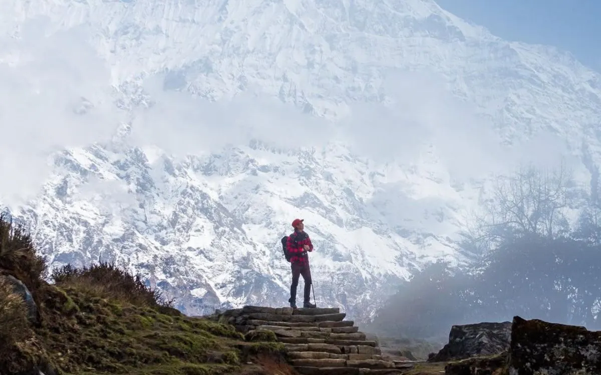 Hiker in red plaid jacket standing on stone steps at Mardi Himal Base Camp with Mount Machhapuchhre (Fishtail) and Annapurna South peaks covered in snow visible through mist
