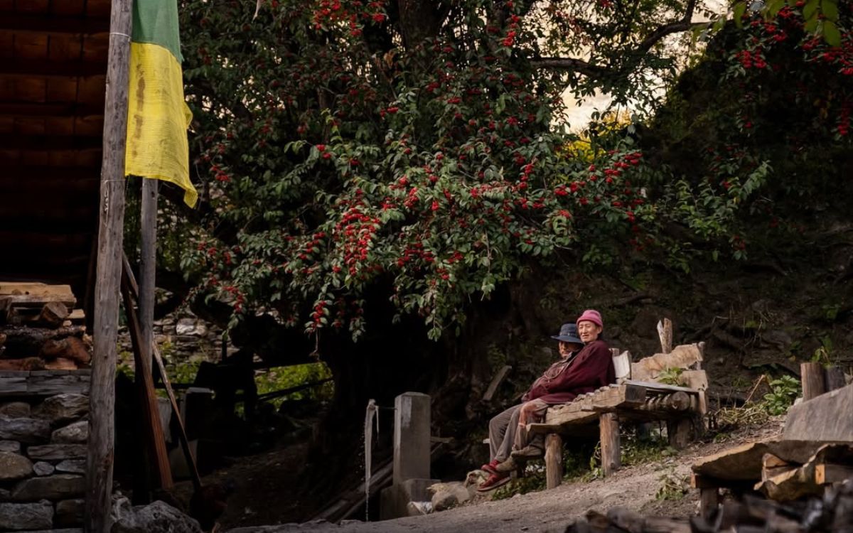 Tibetan Buddhist villagers sitting on wooden bench under prayer flag near stone house Manaslu region Nepal