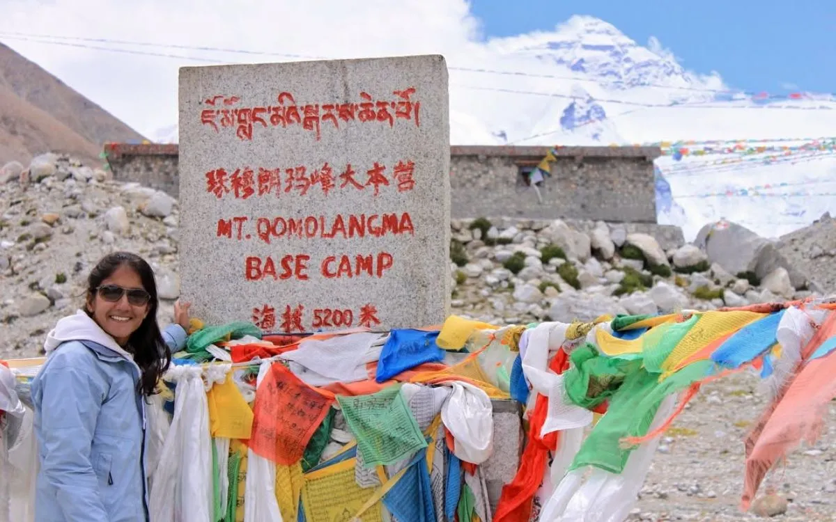 Woman posing next to Mt. Qomolangma Base Camp sign decorated with colorful Tibetan prayer flags