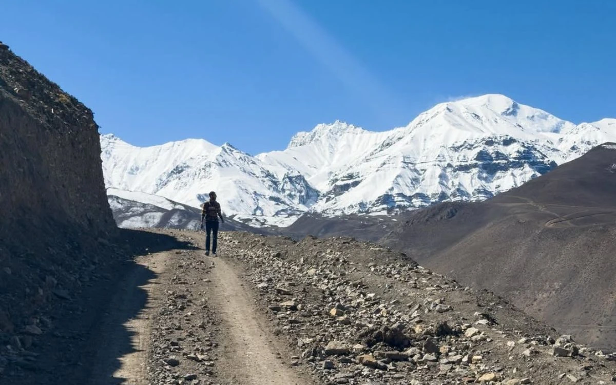 A solo trekker walking a rocky dirt trail toward a massive snow-covered mountain range in Upper Mustang