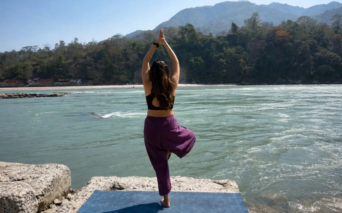 Person doing tree pose yoga exercise on riverbank with mountains in background